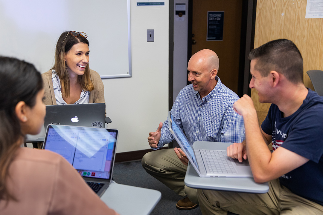 Three students seated at desks in a classroom while a professor crouches at eye level to talk with them
